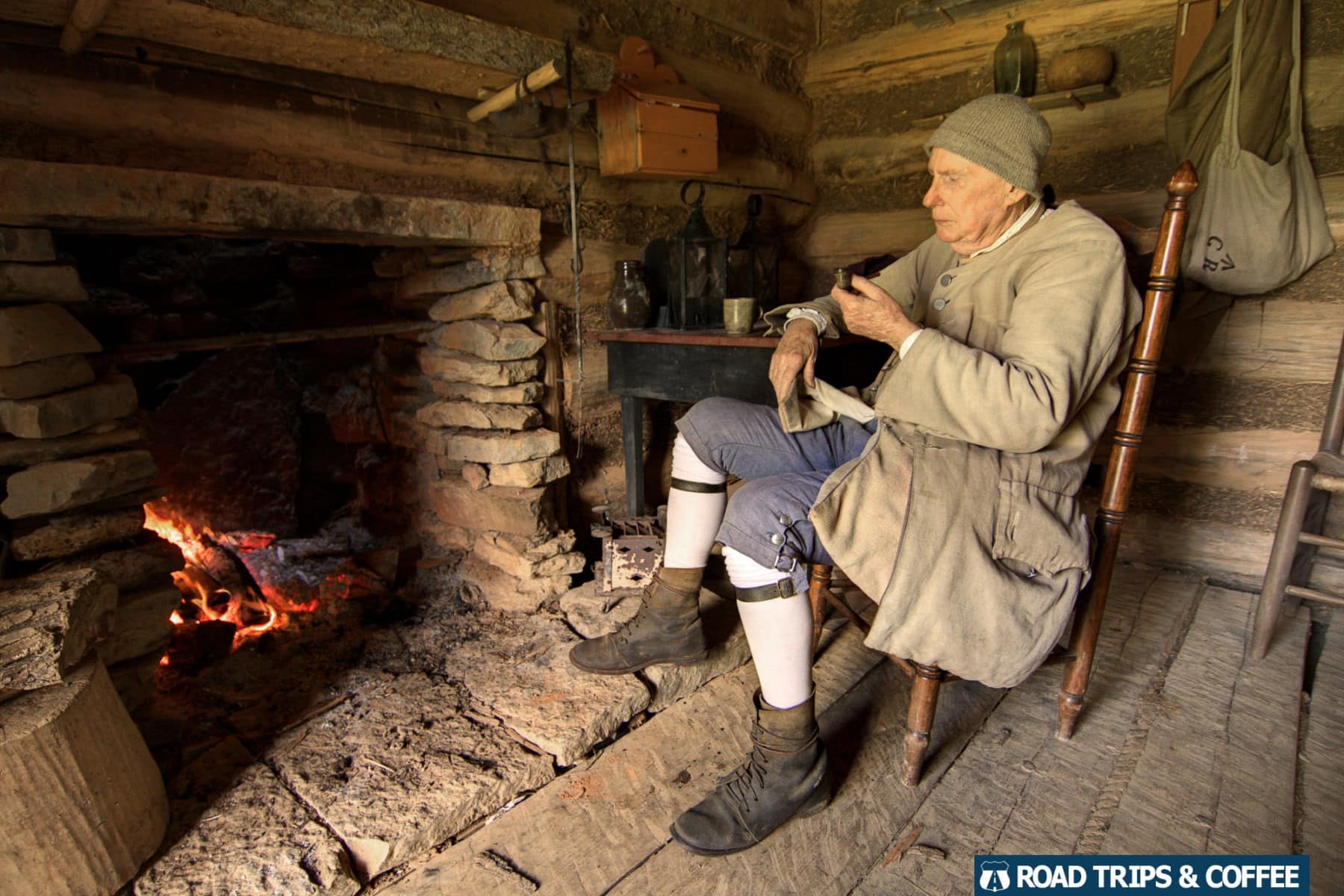 A reenactor dressed in 1700s clothing sitting in front of a fireplace inside a cabin at Martin's Station at Wilderness Road State Park in Virginia
