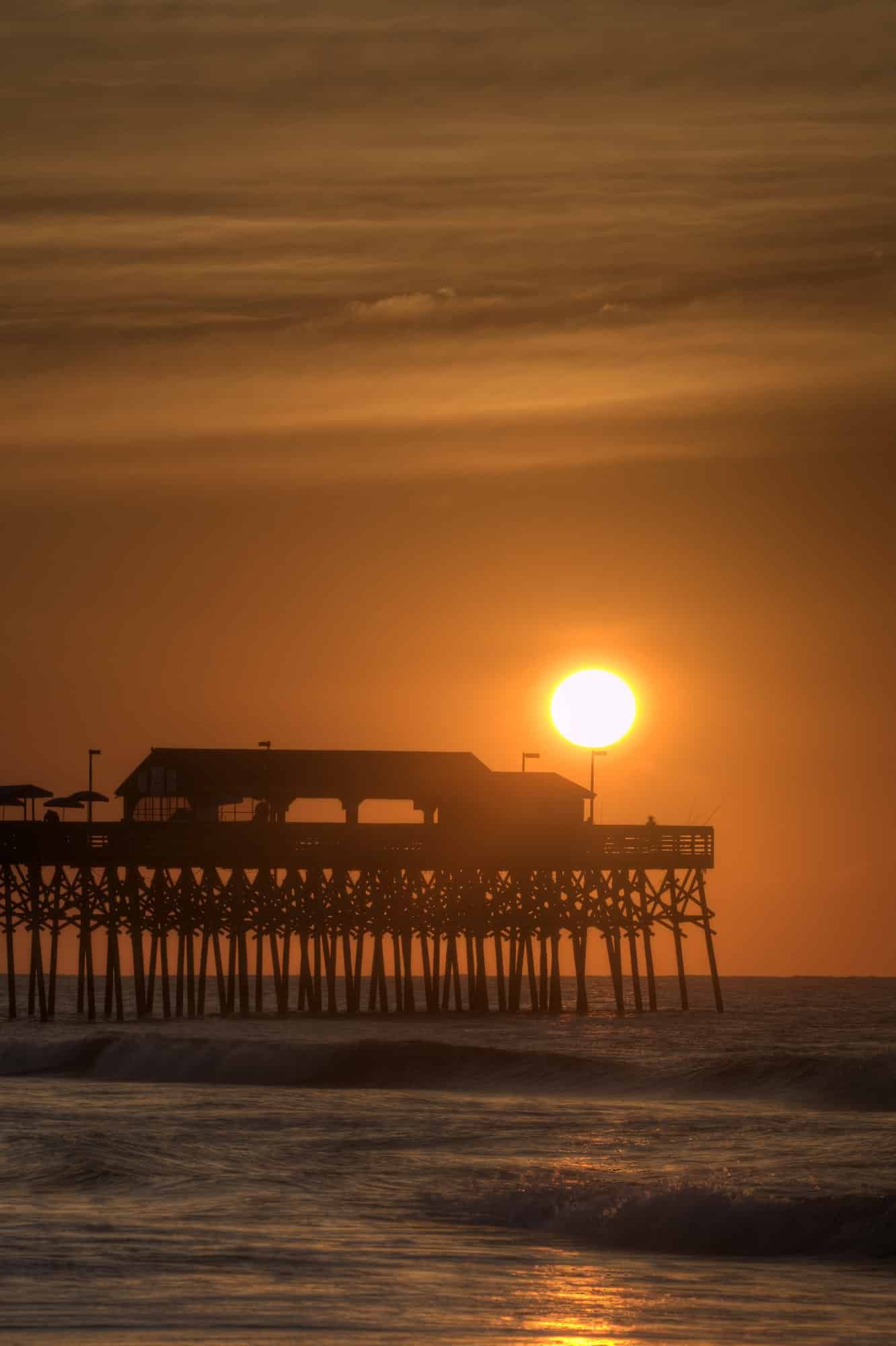 A warm orange sunrise over the black silhouetted Pier at Garden City Beach in South Carolina.