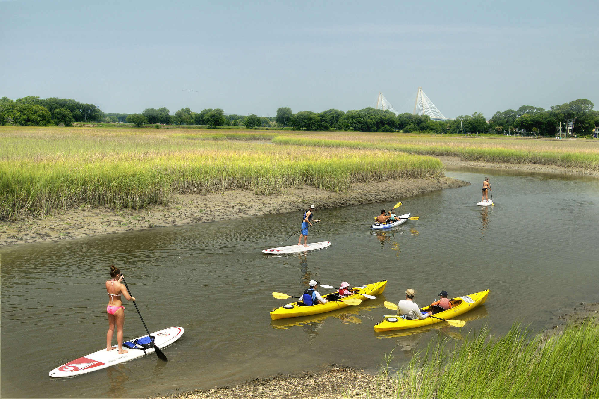 A group of paddleboarders and kayakers move along Shem Creek with a view of the Ravenel Bridge in the distance in Charleston, South Carolina.